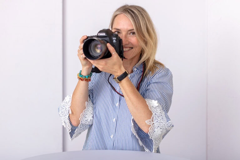 Mel Bogisch steht an einem Tisch und hält einen Fotoapparat halb vor das Gesicht.