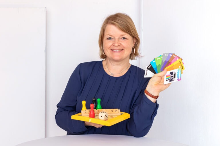 Elke Hayoz steht an einem Tisch, präsentiert diverse Spiele und Karten und lächelt in die Kamera
