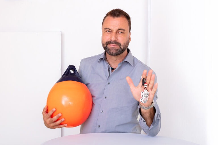 Simon Kramis steht an einem Tisch, lächelt in die Kamera und präsentiert eine Boye und einen Bootsschlüssel